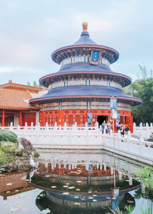 Ornate circular temple with layered blue tiled roofs, red columns, and intricate decorative details reflected in a calm pond with lily pads in the foreground. White stone railings surround the pavilion while visitors walk near the entrance beneath an overcast sky.