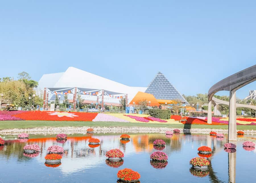 A vibrant flower garden with floating floral arrangements fills the foreground of EPCOT during the Flower and Garden Festival, with the glass pyramids of the Imagination Pavilion and the monorail track in the background under a clear blue sky.