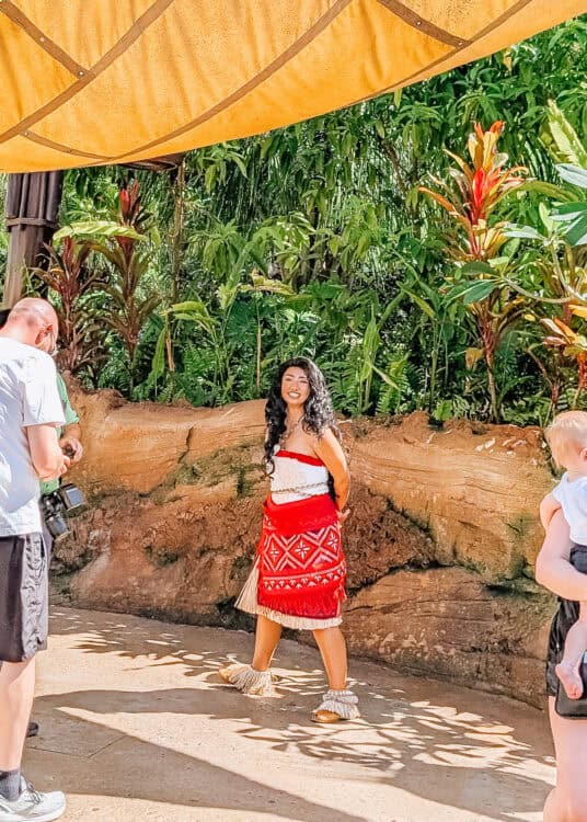 Woman in a red and white Polynesian inspired outfit posing beneath a shaded canopy surrounded by tropical plants and rock walls. Guests stand nearby watching while sunlight filters through the outdoor walkway in the lush theme park setting.