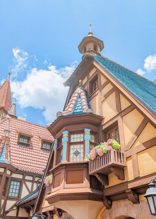 Storybook style building with steep blue rooftops, timber framing, and flower filled balconies set against a bright blue sky with fluffy clouds. Decorative towers and stained glass windows give the architecture a whimsical fairytale appearance.