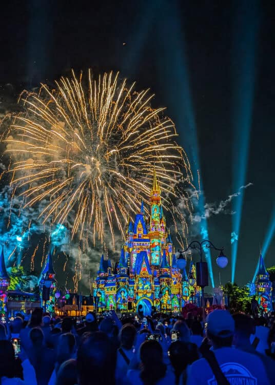 Fireworks burst in the night sky above Cinderella Castle illuminated with colorful character projections while crowds gather below holding up phones to watch the show. Blue beams of light stretch into the sky behind the castle, creating a dramatic nighttime Disney scene.