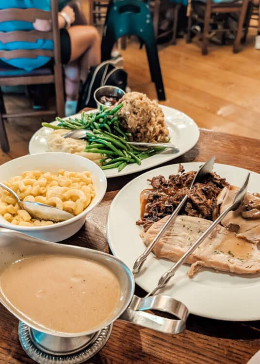 Table filled with comfort food including sliced turkey with gravy, shredded beef, macaroni and cheese, mashed potatoes, green beans, stuffing, and a bowl of creamy soup. Serving utensils rest on the plates while diners sit in the softly lit restaurant background.