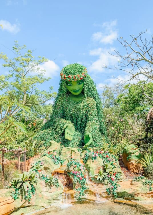 Te Fiti topiary covered in green plants and flowers towers above a small waterfall and tropical garden display at EPCOT. The massive Moana inspired figure sits among rocks and greenery under a bright blue sky, highlighting one of the immersive nature themed attractions in the magic kingdom vs epcot comparison.
