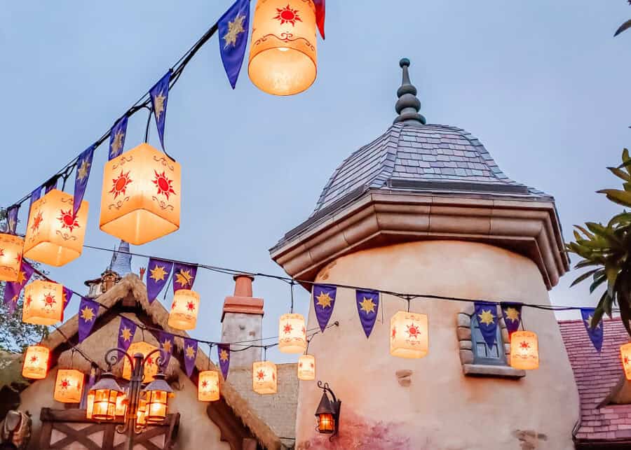 Glowing lanterns decorated with golden sun symbols hang above a fairytale style village with stone towers and shingled rooftops at dusk. Purple banners draped across the walkway add a magical atmosphere to the softly lit fantasy themed setting.