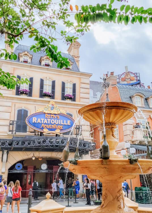 Decorative fountain spraying water in front of the "Remyโs Ratatouille Adventure" attraction sign with Paris inspired buildings and flower boxes in the background. Guests gather beneath the ornate facade while leafy tree branches frame the scene overhead.