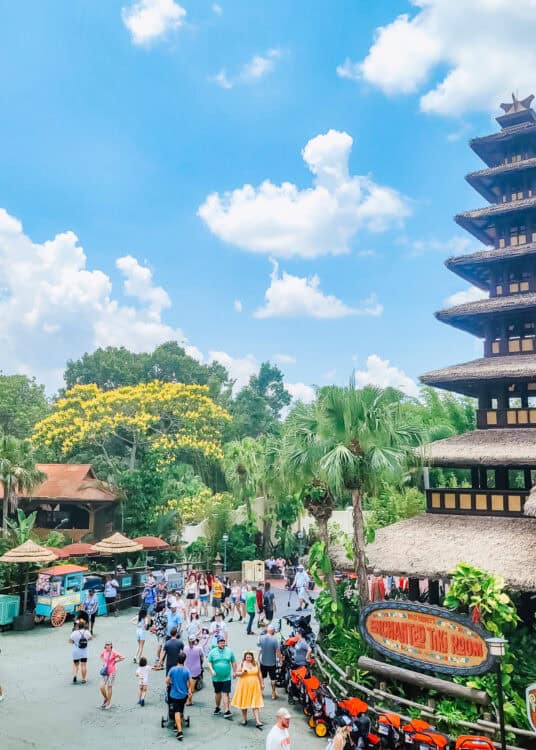 Guests walk through the Adventureland area of Magic Kingdom near Walt Disneyโs Enchanted Tiki Room, with a tall pagoda-style building, tropical plants, and a bright blue sky overhead.