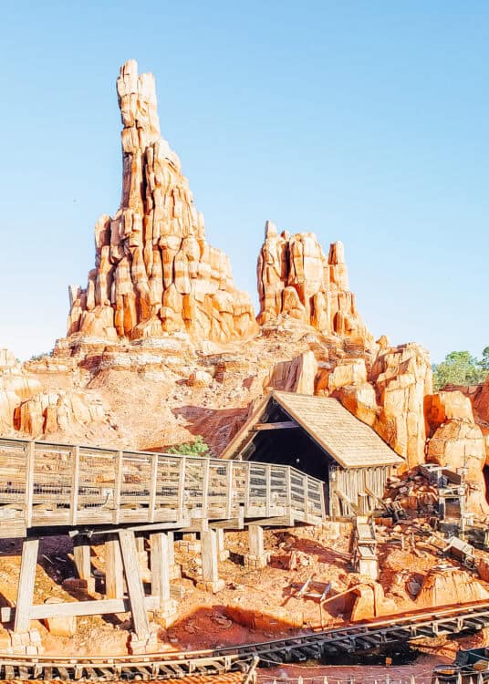 Big Thunder Mountain Railroad rises in towering red rock formations at Magic Kingdom, with a wooden mine tunnel entrance and railroad tracks winding through the desert-style landscape under a clear blue sky.