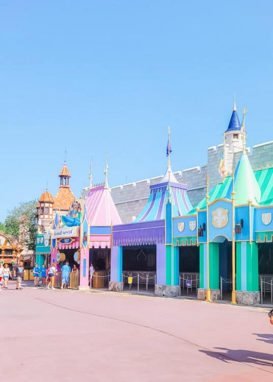 Colorful pastel tents and turrets mark the entrance to the โitโs a small worldโ attraction at Magic Kingdom, with guests walking by under a clear blue sky.