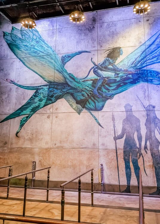 A large mural inside the Avatar Flight of Passage queue at Disneyโs Animal Kingdom shows a Naโvi riding a blue banshee in flight. Overhead lights illuminate the artwork while empty queue rails line the themed interior below.