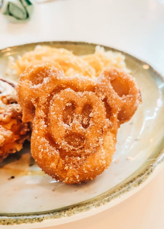 A golden-brown Mickey-shaped waffle dusted with sugar sits on a plate, served with shredded hash browns and breakfast food in the background. The close-up highlights the classic Mickey Mouse shape and crispy texture.