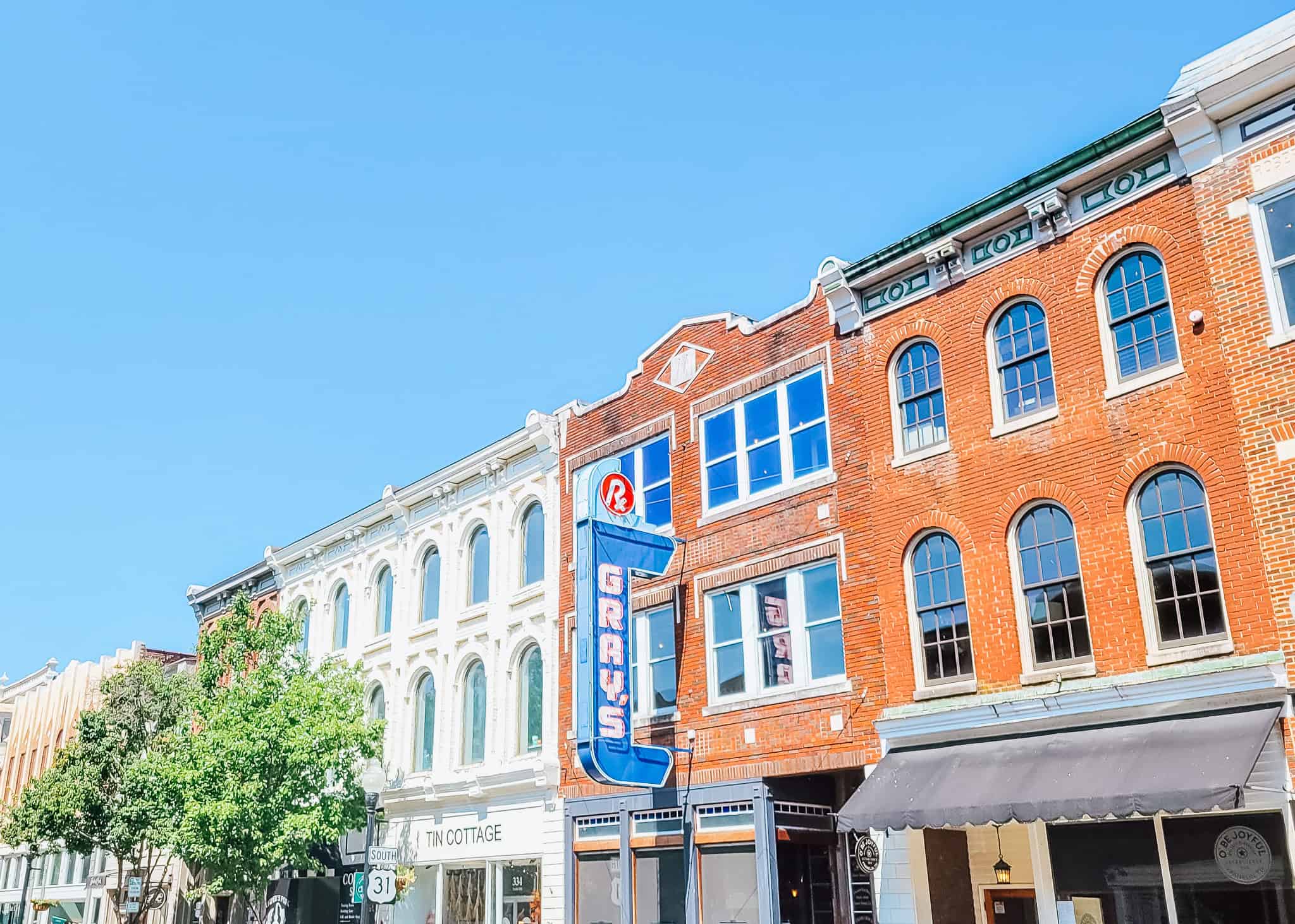 Colorful historic buildings line Main Street in downtown Franklin, Tennessee, with a bright blue “Gray’s” marquee sign mounted on a red brick facade and a white storefront for Tin Cottage nearby. Clear blue skies and green trees add to the inviting small-town charm.