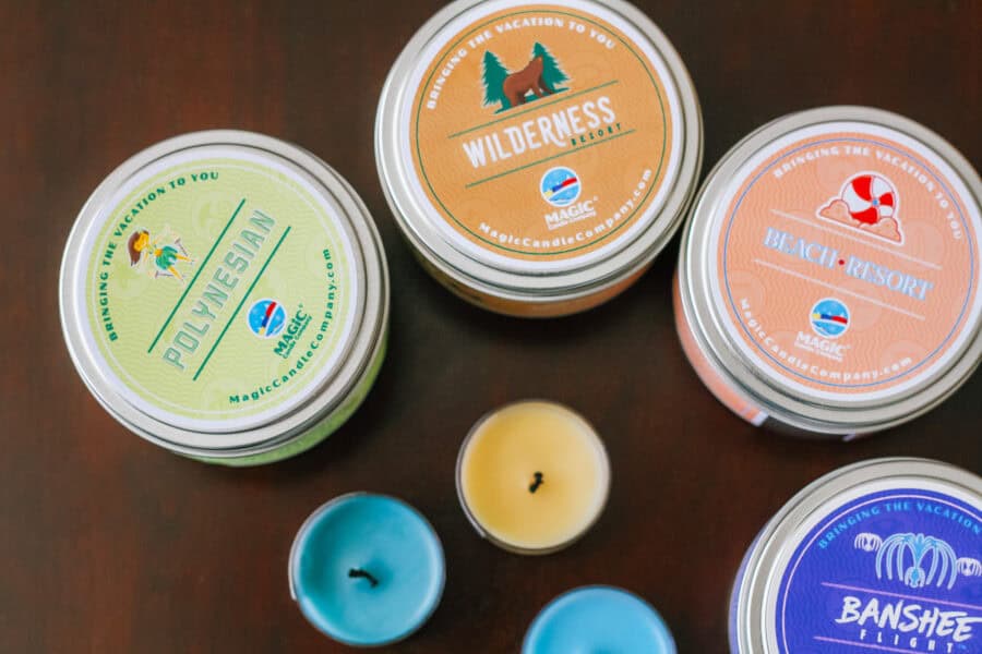 Overhead view of themed candles from Magic Candle Company, including “Polynesian,” “Wilderness Resort,” “Beach Resort,” and “Banshee Flight,” surrounded by small blue and yellow tealight candles on a dark wooden surface.