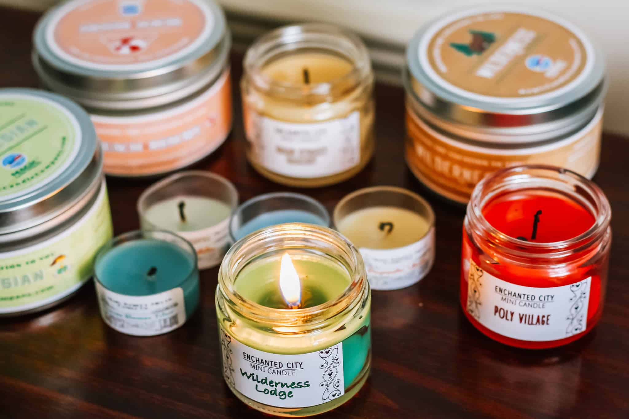 A collection of colorful scented candles in jars and tins displayed on a wooden surface, with one labeled โWilderness Lodgeโ lit in the foreground and others labeled โPoly Village,โ โAmazon,โ and โWildernessโ arranged around it.