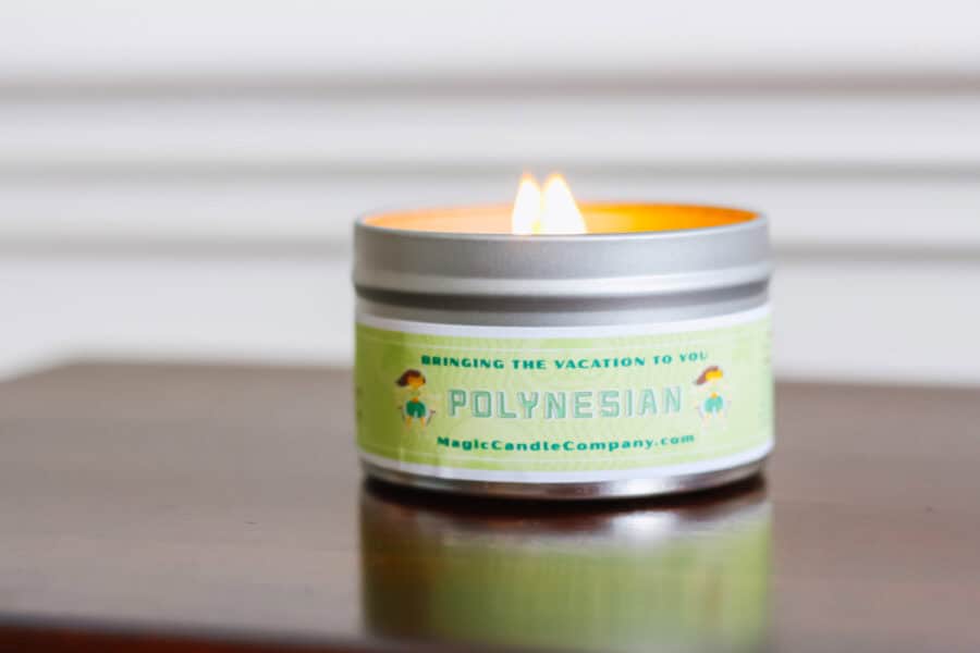 A lit candle in a silver tin labeled “Polynesian” from Magic Candle Company, with a green label that reads “Bringing the Vacation to You,” placed on a wooden surface with a soft white background.
