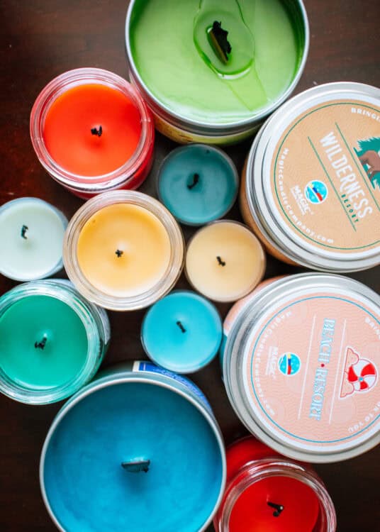 Top-down view of various colorful candles in jars and tins arranged on a wooden surface, featuring labels like “Wilderness Resort” and “Beach Resort” from Magic Candle Company, showcasing a mix of vibrant wax colors including green, red, blue, and yellow.