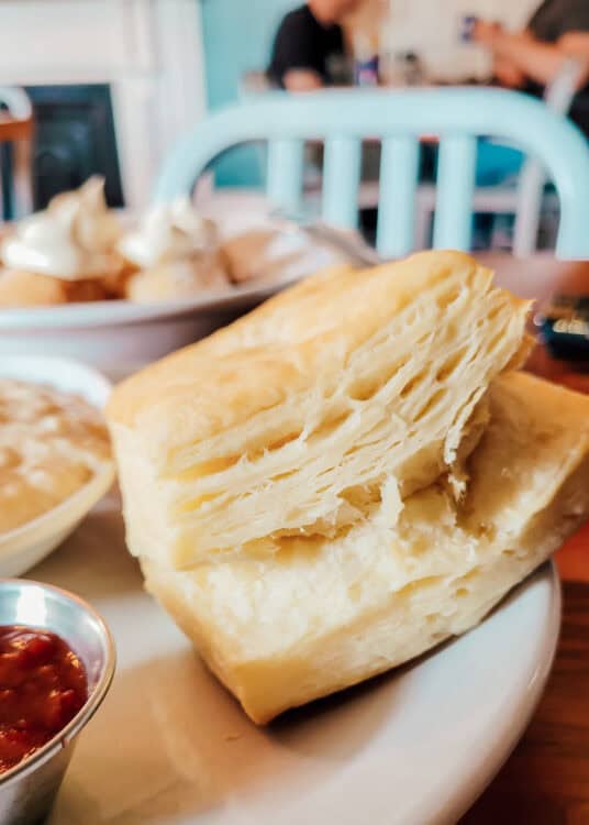 A close-up of a flaky, golden biscuit served on a white plate, accompanied by a side of strawberry jam and a bowl of gravy, with more breakfast items blurred in the background.