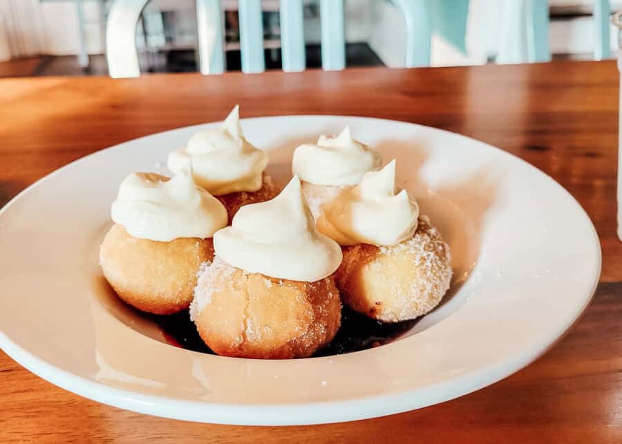 A plate of five round sugar-coated donuts topped with generous swirls of cream, served over a layer of dark berry sauce on a white plate.