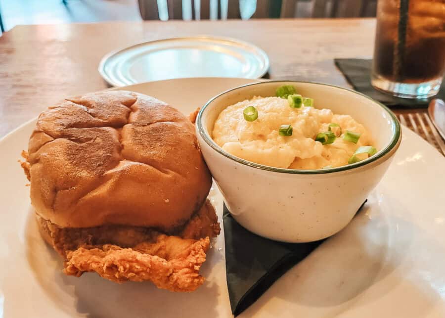 A plate with a crispy fried chicken sandwich on a soft bun, served alongside a bowl of creamy grits topped with chopped green onions.
