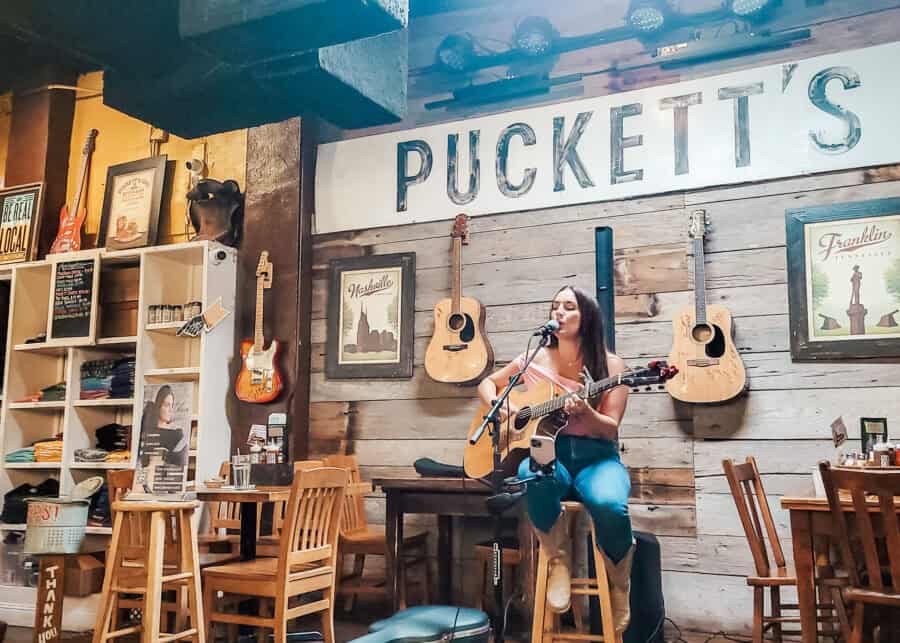 A woman performs live music on an acoustic guitar at Puckett's restaurant in Franklin, Tennessee. She sits on a stool in front of a wooden wall decorated with hanging guitars and framed posters, including ones labeled โNashvilleโ and โFranklin.โ Tables and chairs surround the cozy, rustic space.