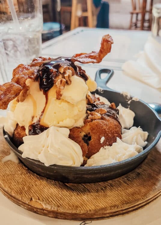 A decadent dessert served in a small cast iron skillet, featuring a warm cookie topped with vanilla ice cream, chocolate sauce, pecans, whipped cream, and crispy bacon strips, presented on a wooden trivet.