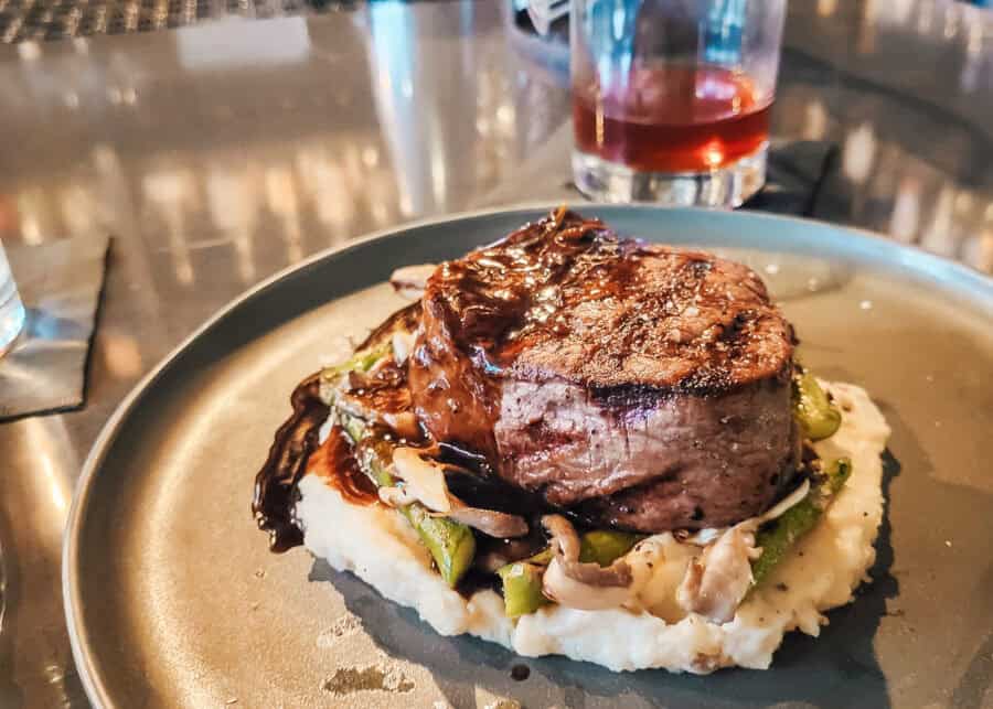 A grilled filet mignon topped with a rich glaze, served on a bed of mashed potatoes, sautรฉed mushrooms, and green vegetables, plated on a matte gray dish with a cocktail in the background.