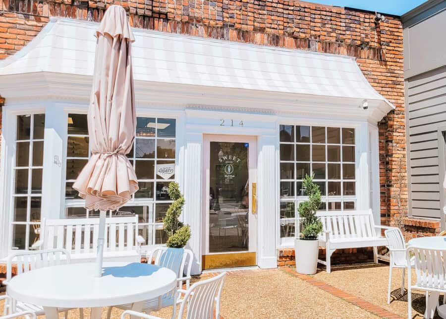 Exterior of a charming brick-front ice cream shop with large windows, white benches, and potted topiary trees. A round white patio table with matching chairs and a closed beige umbrella sits in the foreground under bright sunlight.