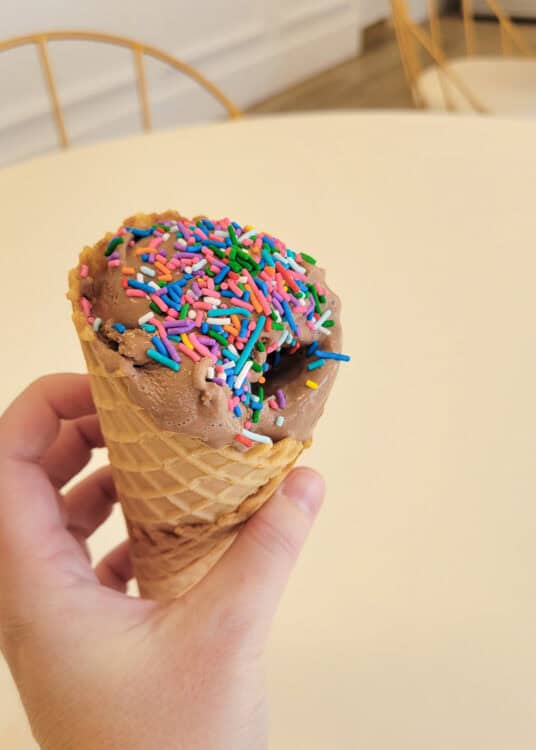 Hand holding a waffle cone filled with chocolate ice cream topped with colorful rainbow sprinkles. The background shows a light-colored table and gold metal chairs in a softly lit cafe setting.