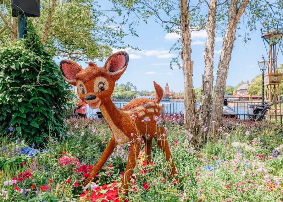 Bambi topiary sculpture made of greenery stands among colorful flowers in a landscaped garden beside a waterfront. Trees and park visitors sit nearby while buildings across the water appear in the background under a bright blue sky.