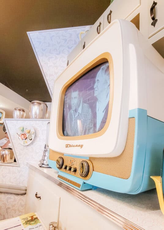 Retro blue and cream television playing a black and white program inside the vintage kitchen at 50s Prime Time Cafe in Disneyโs Hollywood Studios. White cabinets patterned wallpaper and old fashioned kitchenware complete the mid century setting.