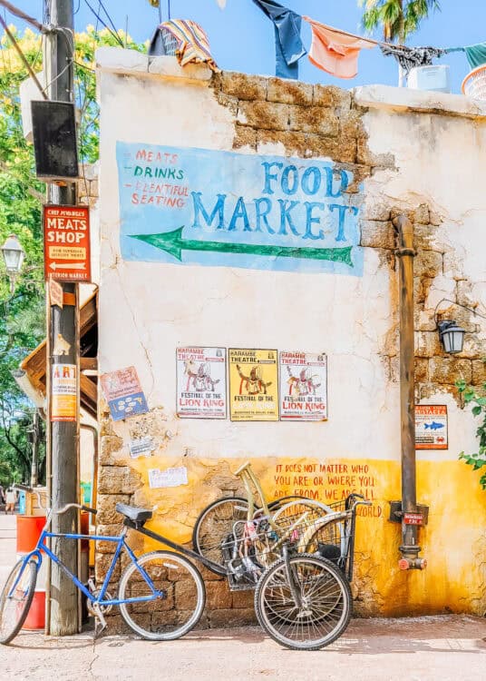 Weathered wall in the Harambe area of Disneys Animal Kingdom painted with the words "FOOD MARKET" and an arrow pointing left alongside signs for "MEATS" "DRINKS" and "PLENTIFUL SEATING". Bicycles lean against the wall beneath colorful Festival of the Lion King posters and other market signs.