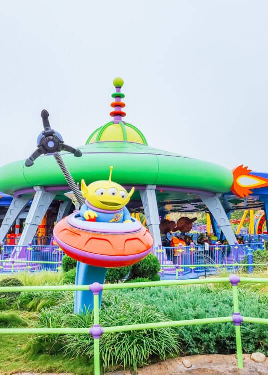 Green three eyed alien from Toy Story sits in a colorful flying saucer outside Alien Swirling Saucers at Disneyโs Hollywood Studios. The bright green and purple attraction canopy and ride vehicles fill the background.