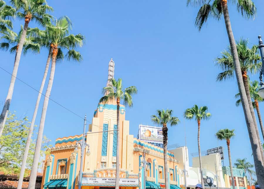 Art Deco buildings and tall palm trees line a sunny street at Disneys Hollywood Studios under a clear blue sky. The colorful architecture features peach turquoise and cream details inspired by classic Hollywood.
