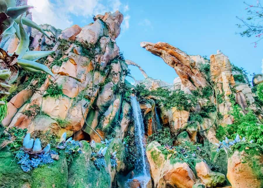 Towering rock formations covered in lush alien plants surround a waterfall in Pandora at Disneys Animal Kingdom. Floating mountain formations rise above the colorful landscape against a bright blue sky.