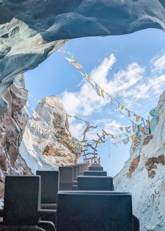 Expedition Everest roller coaster train climbs through a snowy Himalayan mountain scene at Disneyโs Animal Kingdom. Colorful prayer flags stretch overhead against a bright blue sky as riders raise their hands.