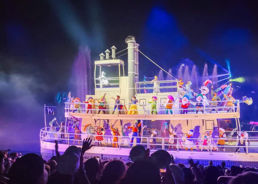 Mickey Mouse Minnie Mouse Donald Duck Goofy Snow White the Seven Dwarfs and other Disney characters perform aboard a brightly lit riverboat at night. Purple fountains and colorful stage lights surround the boat as a large crowd watches the nighttime show.