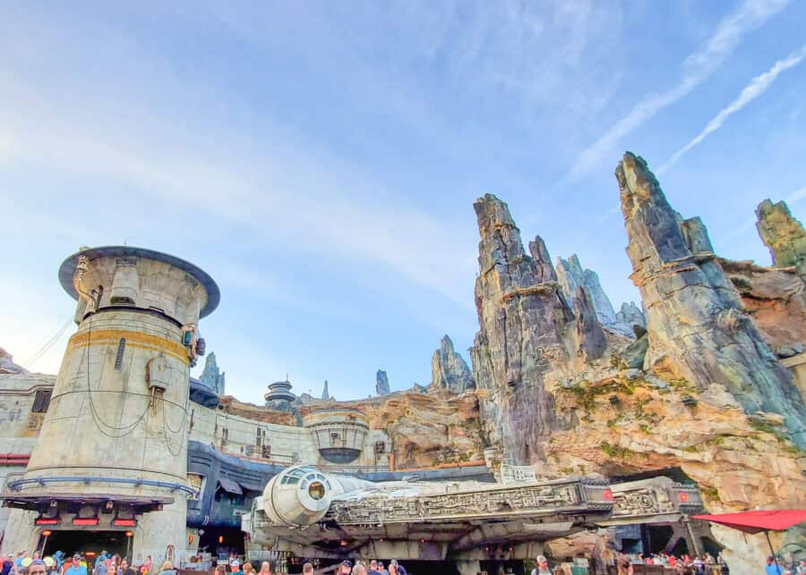 Millennium Falcon parked beneath the towering rocky spires of Star Wars Galaxys Edge at Disneys Hollywood Studios. Weathered futuristic buildings surround the starship as park guests gather below.
