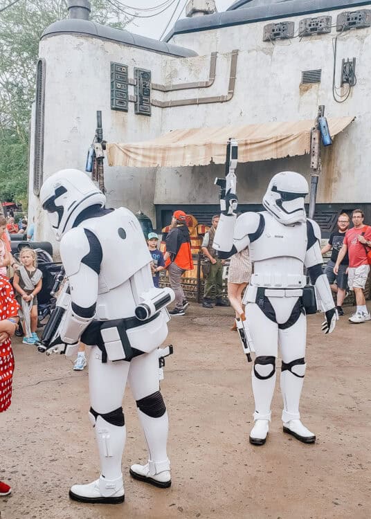 Two First Order Stormtroopers patrol Star Wars Galaxys Edge at Disneyโs Hollywood Studios while carrying blaster props. Guests watch as the Stormtroopers walk through the weathered outpost.