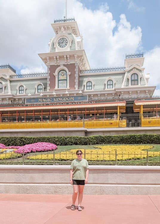 A woman wearing sunglasses, a green T-shirt, and black shorts stands in front of the Magic Kingdom entrance at Walt Disney World. Behind her is the iconic Main Street Train Station with a clock tower, a yellow train, and Mickey Mouse-shaped flower arrangements in vibrant yellow and pink.