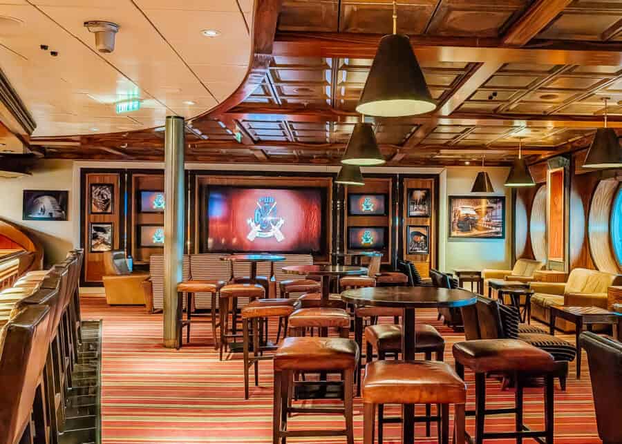 Warm and cozy interior of Pub 687, a Disney Dream restaurant, styled like a classic sports bar with dark wood paneling, leather bar stools, and striped carpeting. A large screen with the Pub 687 logo is framed by sports memorabilia and soft pendant lighting, creating an inviting lounge atmosphere.