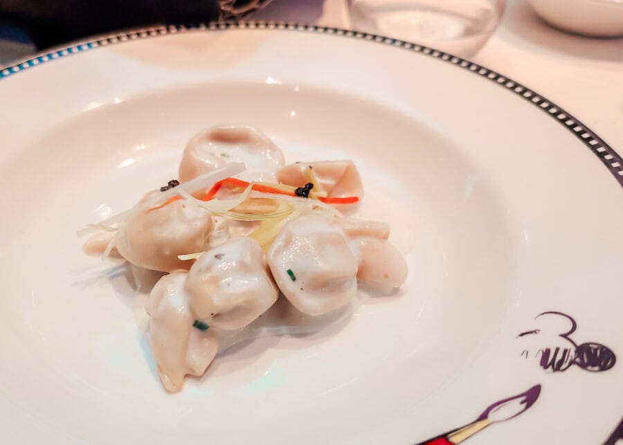 Close-up of a plated pasta dish served at Animator's Palate, featuring plump dumpling-style pasta in a creamy white sauce, garnished with thin julienned vegetables and a small dollop of black caviar. The white plate is decorated with a whimsical Mickey Mouse illustration holding a paintbrush.