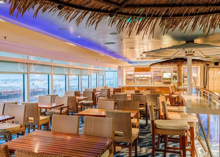 Tropical-themed indoor dining area on a cruise ship, featuring wicker chairs, wooden tables, and thatched-roof details above the bar and seating areas. Large windows line one side of the room, offering a view of the port and docked ships outside.