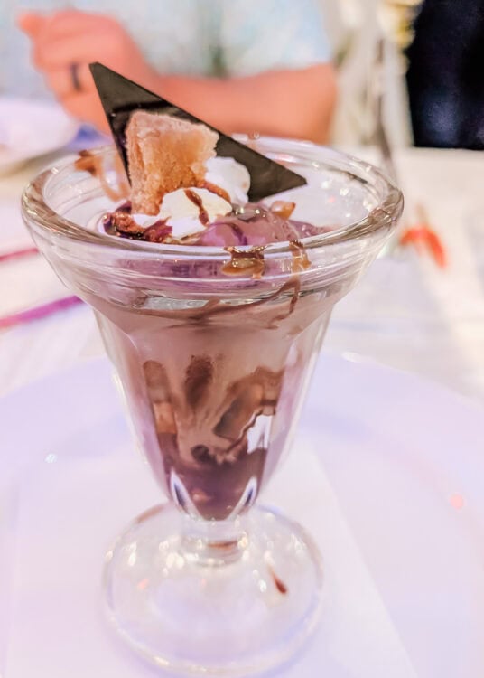 Chocolate sundae served in a tall glass dish, layered with chocolate sauce and topped with whipped cream, a triangle of dark chocolate, and a cookie piece. The dessert is set on a white plate with a napkin underneath.