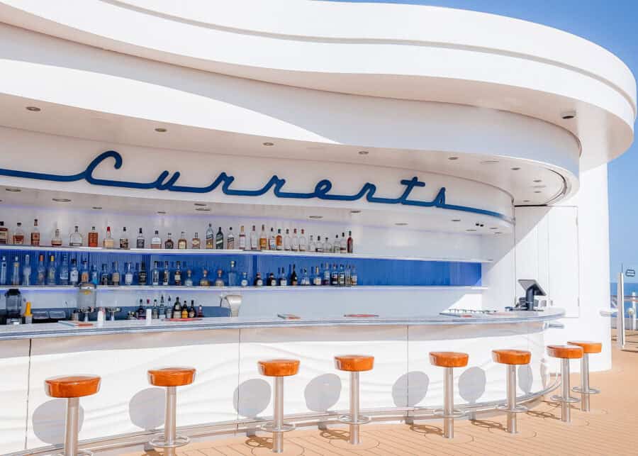 Outdoor bar named "Currents" on a Disney cruise ship, featuring a sleek white and blue design with a wave-like counter, orange bar stools, and a full selection of liquor bottles on display. The bar is set against a clear blue sky, offering a breezy, ocean-view atmosphere.