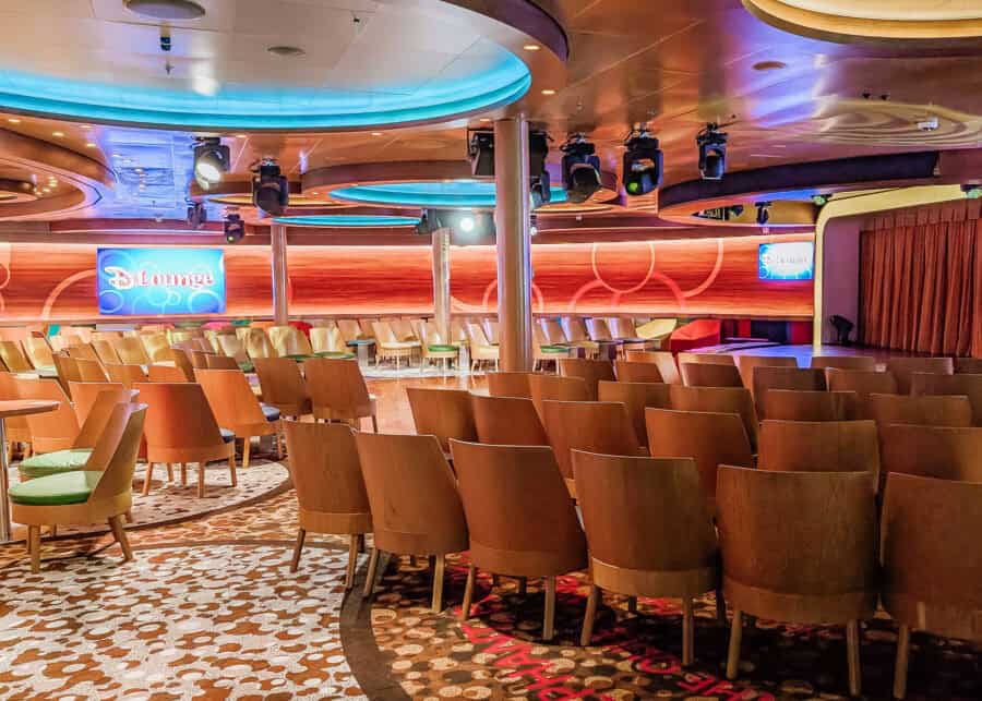 Spacious interior of the D Lounge on a Disney cruise ship, featuring rows of modern wooden chairs, circular stage lighting on the ceiling, and a cozy stage with “D Lounge” displayed on twin wall screens. The room is designed for family entertainment and activities, with vibrant carpeting and colorful accents.