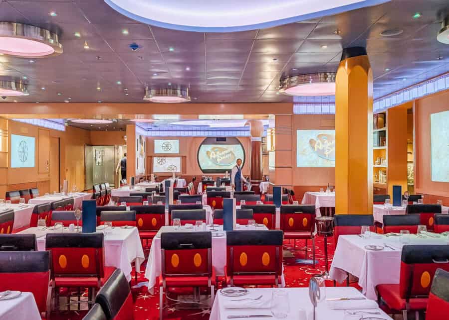 Bright and colorful dining room at Animator's Palate on a Disney cruise ship, featuring red chairs with yellow buttons styled like Mickey Mouse, white tablecloths, and animated screens on the walls. A server in a white shirt and black vest walks between tables under soft purple ceiling lights.