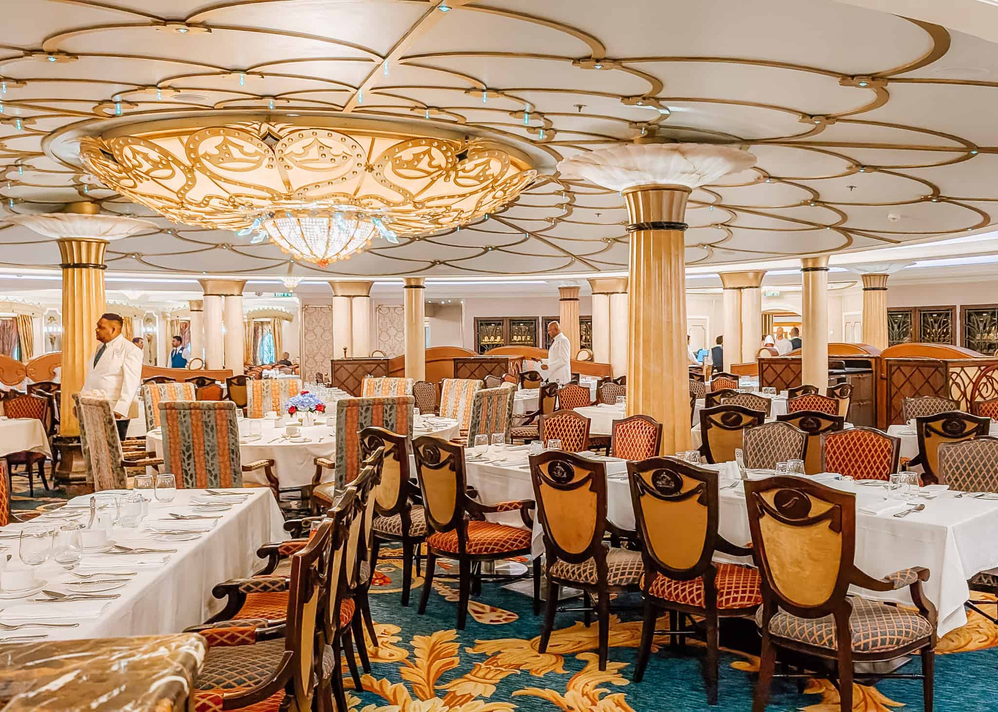 Opulent dining room on a Disney cruise ship, adorned with an intricate golden chandelier, white-clothed tables, and a variety of richly upholstered chairs. The space is accented by large golden columns and a vibrant carpet with ornate leaf designs, while servers prepare for dinner service.