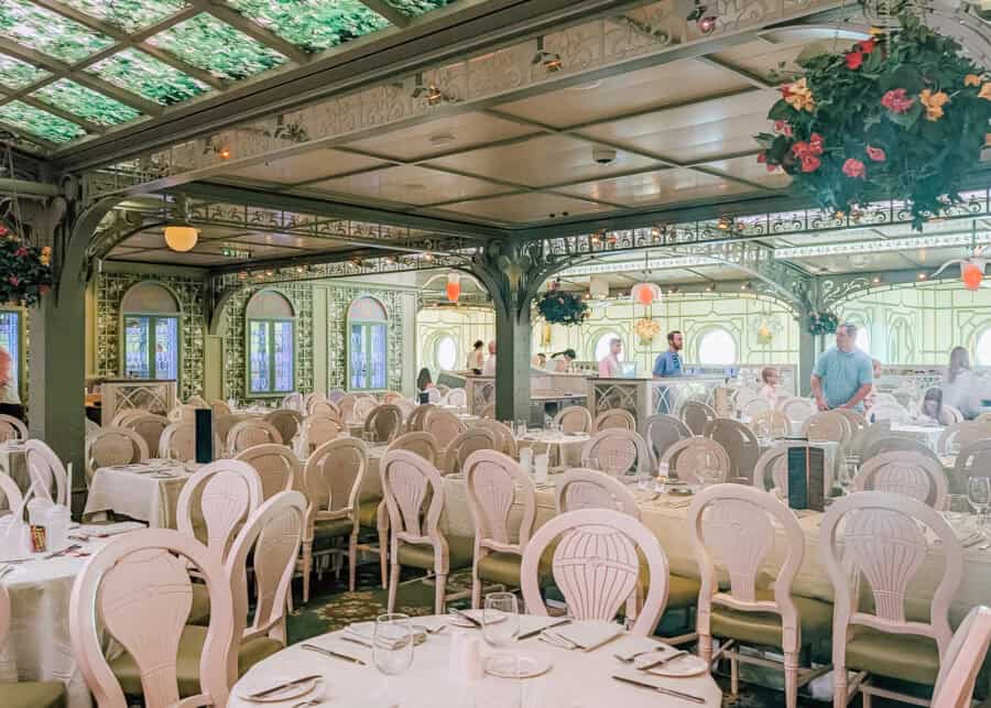 Elegant dining room aboard a Disney cruise ship, decorated in a garden-inspired style with white carved chairs, hanging flower baskets, and green stained-glass ceiling panels. Soft natural light filters through arched windows, while guests begin to arrive and take their seats.