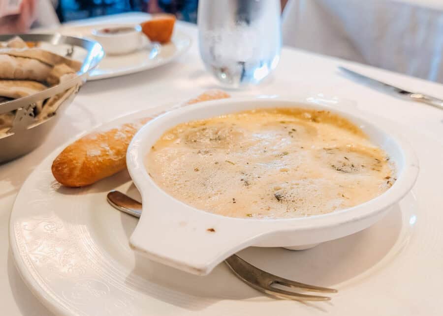 Bubbling dish of escargot baked in garlic herb butter, served in a white ceramic dish alongside a golden-brown breadstick. The dish is plated on a white tablecloth with a seafood fork resting beside it, ready for dining.