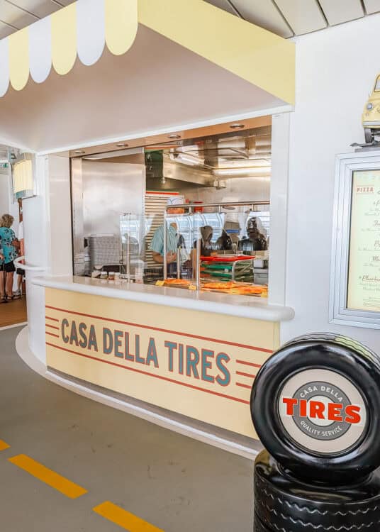 Pizza counter themed as "Casa Della Tires" from Disney Pixar’s Cars, with a retro yellow and red design, a chef behind the glass preparing fresh pizzas, and a stacked tire display labeled "Quality Service" on the side. The playful setup mimics a pit stop, blending automotive decor with a casual dining experience.