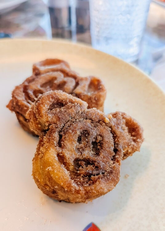 Close-up of two Mickey Mouse-shaped cinnamon rolls coated in sugar, with visible swirls of cinnamon and a golden-brown crust. The pastries are served on a cream-colored plate with a water glass in the background.